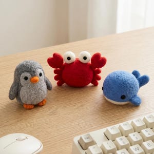 Pode incluir: Três figuras de animais de feltro: um pinguim cinzento com bico e pés laranja, um caranguejo vermelho e uma baleia azul. As figuras estão sobre uma superfície de madeira castanha clara, perto de um rato e um teclado de computador.