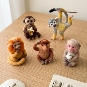 May include: A collection of five handcrafted felt monkey figurines in various poses. One holds a banana, another is hanging from a stick, and others sit or pose. The monkeys are brown, yellow, gray, and orange, set on a wooden desk.