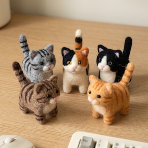 May include: Five handcrafted felt cat figurines in various colours and patterns, including grey tabby, brown tabby, calico, black and white, and orange tabby. The cat figurines are standing on a light brown wooden surface.