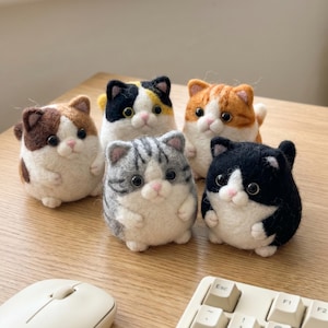 May include: Six felt cat figurines in assorted colours, including brown, black, orange, and grey, with white details. The cat toys are arranged on a wooden surface, with a computer mouse and keyboard in the foreground.