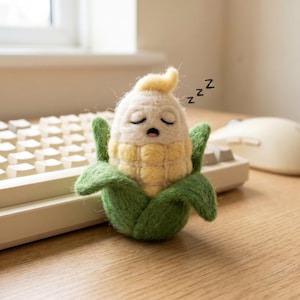 May include: A needle-felted corn figurine with a cream-coloured top and yellow kernels, wrapped in green husks. The corn has a sleeping expression with closed eyes, an open mouth, and three black 'z' characters floating to its right side.