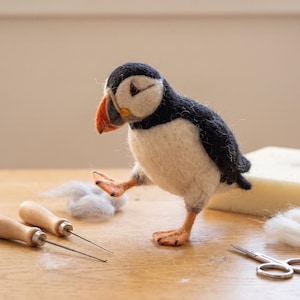 May include: A needle-felted puffin figurine with a black back, white chest, and a bright orange beak. The bird stands on orange feet with one leg raised. Two wooden-handled felting needles, loose wool fibres, and metal scissors are nearby.