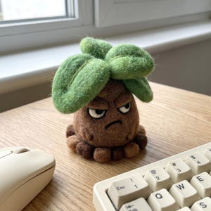 May include: A handmade felted figure of a grumpy-faced plant with green leaves and a brown body. The figure is sitting on a wooden desk next to a computer mouse and keyboard. The figure is approximately 4 inches tall.