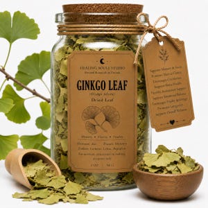 May include: A clear glass jar filled with dried ginkgo leaves, topped with a cork stopper. The label reads HEALING SOULS STUDIO GINKGO LEAF, 2 OZ, 56 G. A brown tag lists benefits, and a wooden scoop and small bowl contain loose dried leaves.
