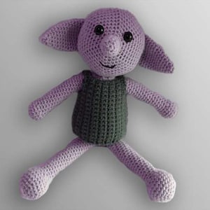 May include: A handmade crochet doll in shades of purple and gray. The doll has large ears, a long nose, and black eyes. It is wearing a gray vest. The arms and legs are outstretched, giving the doll a playful appearance.