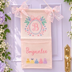 May include: A light pink fabric wall hanging with a pocket, featuring an embroidered letter B inside a floral wreath, the name Brynnlee, and a row of five colorful dress icons. The piece is accented with two sheer pink bows at the top.