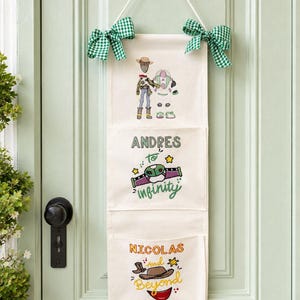 May include: A vertical cream-coloured fabric wall organiser with three pockets, featuring embroidered Toy Story characters and the text "ANDRES TO INFINITY" and "NICOLAS and Beyond". It is hung by a white cord with green gingham bows on each side.