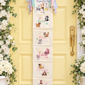 May include: A vertical off-white fabric wall organizer with six pockets, featuring embroidered names and colorful cartoon animal illustrations. The top pocket reads Kugma Family, followed by Kevin, Renee, Lilian, Caroline, and Madeline.