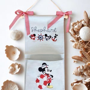 May include: A light gray fabric wall hanging organizer with a pocket, featuring the embroidered name Shephard, nautical icons, and a sailor-themed mouse character holding a life preserver. The top is accented with two red and white gingham bows.