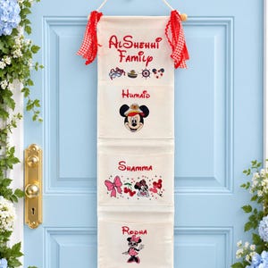 Op de afbeelding: Een crèmekleurige stoffen wandhanger met drie vakken, voorzien van rode tekst en cartoonafbeeldingen. Bovenaan staat AlShehhi Family, gevolgd door vakken met de namen Humaid, Shamma en Rodha, versierd met nautische motieven en strikken.