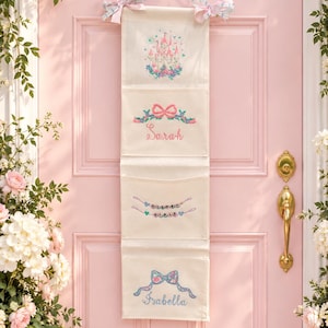 May include: A vertical cream-colored fabric wall organizer with four pockets, featuring embroidered designs including a pink castle, a bow with the name Sarah, a string of beads spelling DISNEY GIRLS, and a bow with the name Isabella.