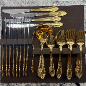 May include: A gold-toned flatware set featuring ornate, embossed handles on knives, spoons, and forks. The collection includes several knives with silver-colored blades, multiple spoons of varying sizes, and forks, all arranged on a dark brown tray.