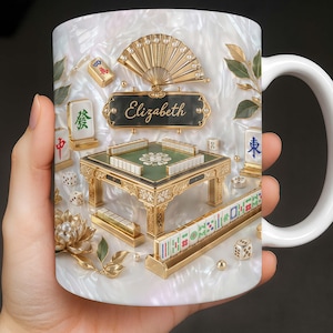 May include: White ceramic mug featuring a detailed, ornate design of a Mahjong set. The design includes a miniature table, tiles, dice, and a fan, all in gold and green. The name "Elizabeth" is displayed on a black plaque.
