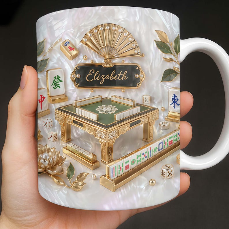May include: White ceramic mug featuring a detailed, ornate design of a Mahjong set. The design includes a miniature table, tiles, dice, and a fan, all in gold and green. The name "Elizabeth" is displayed on a black plaque.