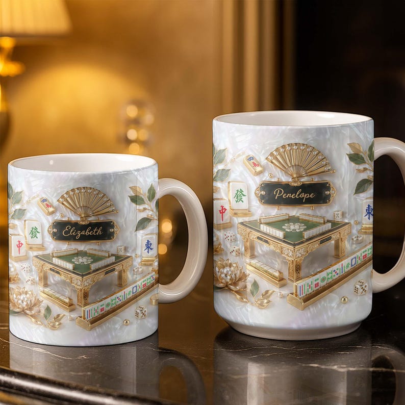 May include: Two white ceramic mugs with a pearlized finish, featuring a detailed design of a Mahjong table, fan, and tiles. One mug has the name "Elizabeth" and the other "Penelope". The mugs have gold accents and cream-colored handles.