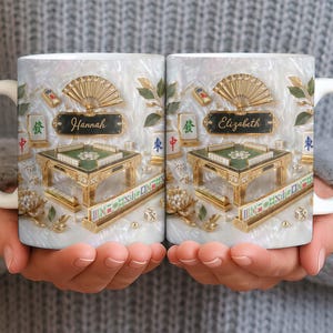May include: White ceramic mug featuring a detailed Mahjong design with gold accents, a fan, and the name "Hannah" on one side and "Elizabeth" on the other. The design includes a miniature table, tiles, and dice, set against a pearlescent background.
