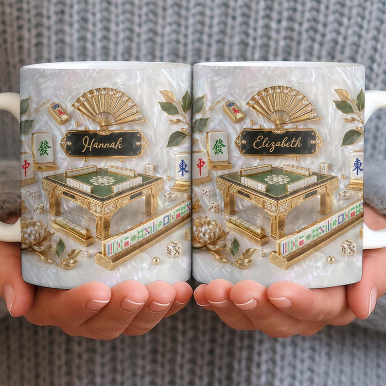 May include: White ceramic mug featuring a detailed Mahjong design with gold accents, a fan, and the name "Hannah" on one side and "Elizabeth" on the other. The design includes a miniature table, tiles, and dice, set against a pearlescent background.