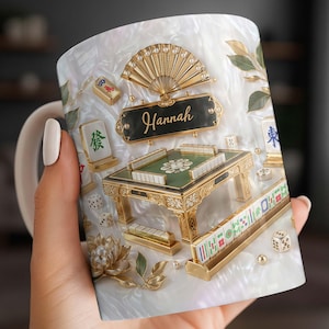 May include: White ceramic mug featuring a detailed design of a Mahjong table, tiles, dice, and a fan. The mug has a pearlescent background and the name "Hannah" is displayed on a black plaque. Gold accents enhance the design.