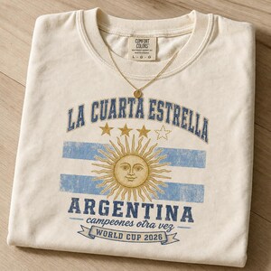 May include: A cream-colored crewneck t-shirt featuring a graphic with the text LA CUARTA ESTRELLA, ARGENTINA, campeones otra vez, and WORLD CUP 2026. The design includes a golden sun with a face, three stars, and blue horizontal stripes.