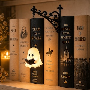 May include: A black metal wall bracket with a scrollwork design holds a small, glowing white ghost figurine suspended by a chain. The ghost is shaped like a sheet with two black eyes and holds a small black book in its hands, positioned between several hardcover books on a shelf.