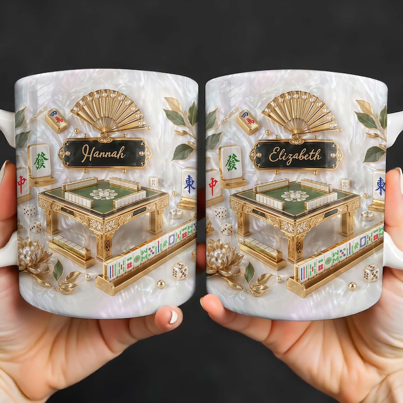 May include: White ceramic mug featuring a detailed design of a Mahjong table with gold accents, dice, and tiles. The mug has a pearlescent background with the name "Hannah" on one side and "Elizabeth" on the other. The design includes a fan and floral elements.