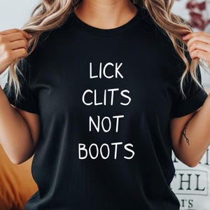 May include: A black short-sleeved t-shirt featuring the white text LICK CLITS NOT BOOTS printed in a simple sans-serif font across the center of the chest. The garment has a crew neckline and a standard fit, showcasing the bold white lettering clearly.