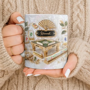 May include: A white ceramic mug featuring a detailed design of a Mahjong set. The design includes a miniature table, tiles, dice, and a fan, with the name "Hannah" displayed on a black plaque. The mug is held by a person wearing a beige sweater.