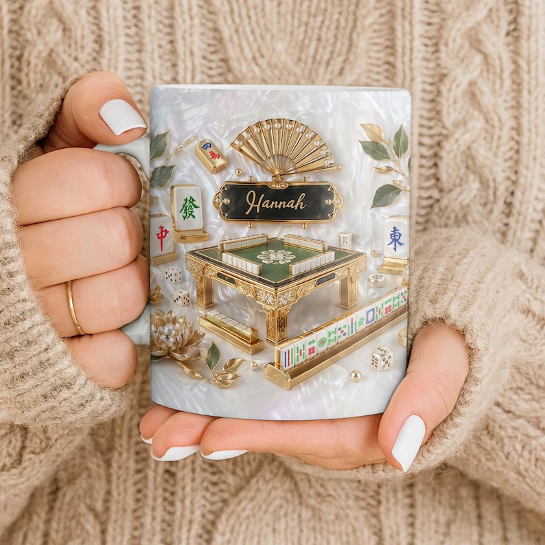 May include: A white ceramic mug featuring a detailed design of a Mahjong set. The design includes a miniature table, tiles, dice, and a fan, with the name "Hannah" displayed on a black plaque. The mug is held by a person wearing a beige sweater.