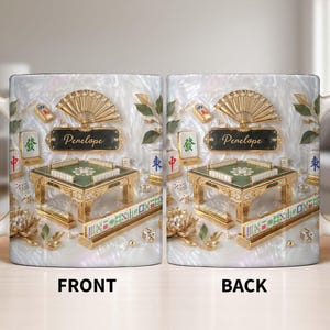 May include: A white ceramic mug featuring a detailed illustration of a Mahjong table, dice, and tiles. The design includes a gold fan, floral accents, and the name "Penelope" on a black plaque. The front and back of the mug are shown.