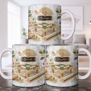 May include: White ceramic mug featuring a detailed design of a Mahjong table, tiles, and decorative elements. The mug has a gold and green color scheme with the name "Penelope" on a black plaque. The design includes a fan and floral accents.