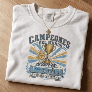 May include: A white crew neck t-shirt featuring a graphic print with the text CAMPEONES DEL MUNDO, otra vez ARGENTINA, and WORLD CUP 2026. The design includes a gold trophy, a sun emblem, and blue and yellow rays. A gold necklace rests on the collar.