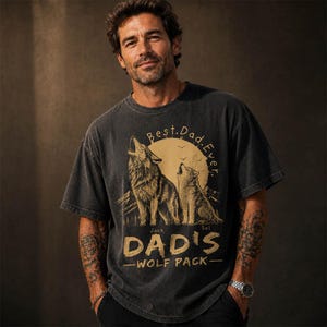 May include: Dark gray t-shirt with a graphic of two wolves howling at a full moon, mountains, and the text "Best. Dad. Ever." and "Dad's Wolf Pack." The shirt is worn by a person.