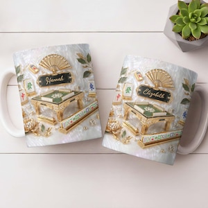 May include: Two white ceramic mugs with a detailed design featuring a golden mahjong table, fan, and tiles. One mug has "Hannah" and the other "Elizabeth" on a black nameplate. The background has a pearlescent effect.