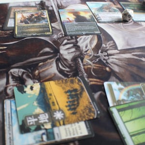 May include: A tabletop scene featuring various trading card game cards, a blue die, and a six-sided die. The cards display fantasy art and text. The background is a playmat with a detailed illustration.