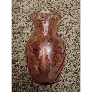 May include: A pink glass vase with a swirling pattern. The vase has a wide base that tapers to a narrower neck and flares out at the top. The glass has a textured appearance, and the color is a muted pink.