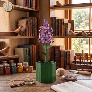Könnte beinhalten: Eine dekorative Metallblume mit lila Blütenblättern und einem grünen Stiel, platziert in einem grünen sechseckigen Topf. Die Blume steht auf einem Holztisch mit Büchern, Gläsern und einer Lupe. Der Hintergrund umfasst Bücherregale und Holzvogelfiguren.