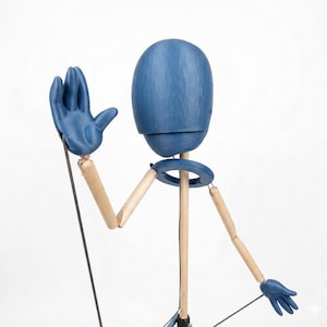 May include: A blue wooden puppet with articulated limbs and a round head. The puppet's hands and head are painted blue, while the arms and legs are made of natural wood. The puppet is posed with one hand raised in a waving gesture.