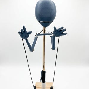 May include: A blue marionette figure with a smooth, rounded head and articulated arms, suspended by black strings from a wooden control bar. The figure features a central wooden support rod attached to a square wooden base, with black handles.