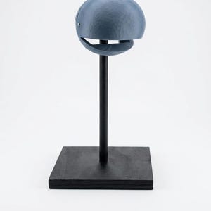 Puede incluir: Un objeto esférico azul texturizado con una abertura de ranura horizontal, montado sobre una varilla vertical negra delgada unida a una base cuadrada negra. La esfera presenta una textura de grano de madera visible y un pequeño cierre metálico.