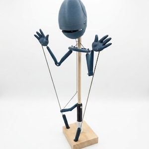 Könnte beinhalten: Eine blaue Marionette mit einem ovalen Kopf, beweglichen Gliedmaßen und fünf-fingrigen Händen. Die Figur ist an einem zentralen Holzstab befestigt, der auf einem quadratischen Holzsockel steht, mit dünnen Kontrollschnüren an Armen und Beinen.