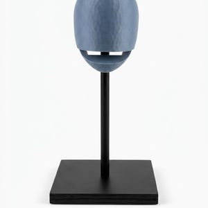 May include: A blue, egg-shaped object with a textured, faceted surface and a horizontal opening, mounted on a thin black vertical rod attached to a square black base. The object is positioned centrally, showcasing a smooth, matte finish.
