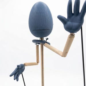 May include: A whimsical wooden puppet with a blue egg-shaped head and blue hands. The puppet's arms and legs are made of light-colored wood, with joints allowing for movement. The puppet is posed with one hand raised in a greeting gesture.