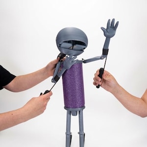 Puede incluir: Una marioneta articulada gris con cabeza esférica de boca ancha, torso cilíndrico de espuma morada y extremidades grises delgadas. La marioneta cuenta con varillas de control negras unidas a los brazos y la cabeza para su manipulación.