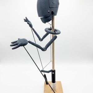 Puede incluir: Una figura articulada gris con una cabeza lisa y redondeada y extremidades articuladas, montada sobre un poste de madera vertical unido a una base cuadrada de madera. La figura tiene finos hilos de control negros conectados a las manos y al torso.