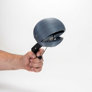 May include: A handheld waste scooper featuring a matte grey, spherical, hinged head attached to a black plastic handle. The mechanism includes a spring-loaded trigger and a thin black cord, designed for picking up items with a clamping motion.