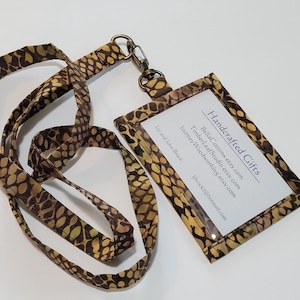 May include: A handcrafted lanyard with a brown and yellow patterned design. The lanyard has a clear plastic ID holder with the text "Handcrafted Gifts" and several Etsy shop URLs.
