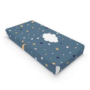 Könnte beinhalten: Ein Spannbettlaken für das Kinderbett mit einem Nachthimmel-Design. Das Laken ist dunkelblau mit einem Muster aus weißen Wolken, gelben Sternen und kleinen Punkten in Weiß, Rosa und Marineblau. Ein gelber Halbmond ist ebenfalls abgebildet.