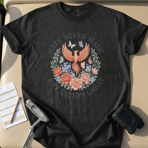 Può includere: T-shirt nero con un design floreale con una fenice, farfalle e la scritta "Hate Never Made Any Nation Great". Il design include fiori arancioni, rosa e blu. La maglietta è su un campo da golf.
