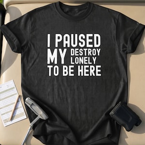 May include: Black t-shirt with the white text "I PAUSED MY DESTROY LONELY TO BE HERE." A golf putter, scorecard, and rangefinder are visible in the image. The shirt is likely made of cotton or a similar material.