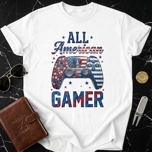 May include: White t-shirt featuring a patriotic design with a video game controller. The controller is decorated with red, white, and blue patterns, the American flag, and the words "ALL AMERICAN GAMER".